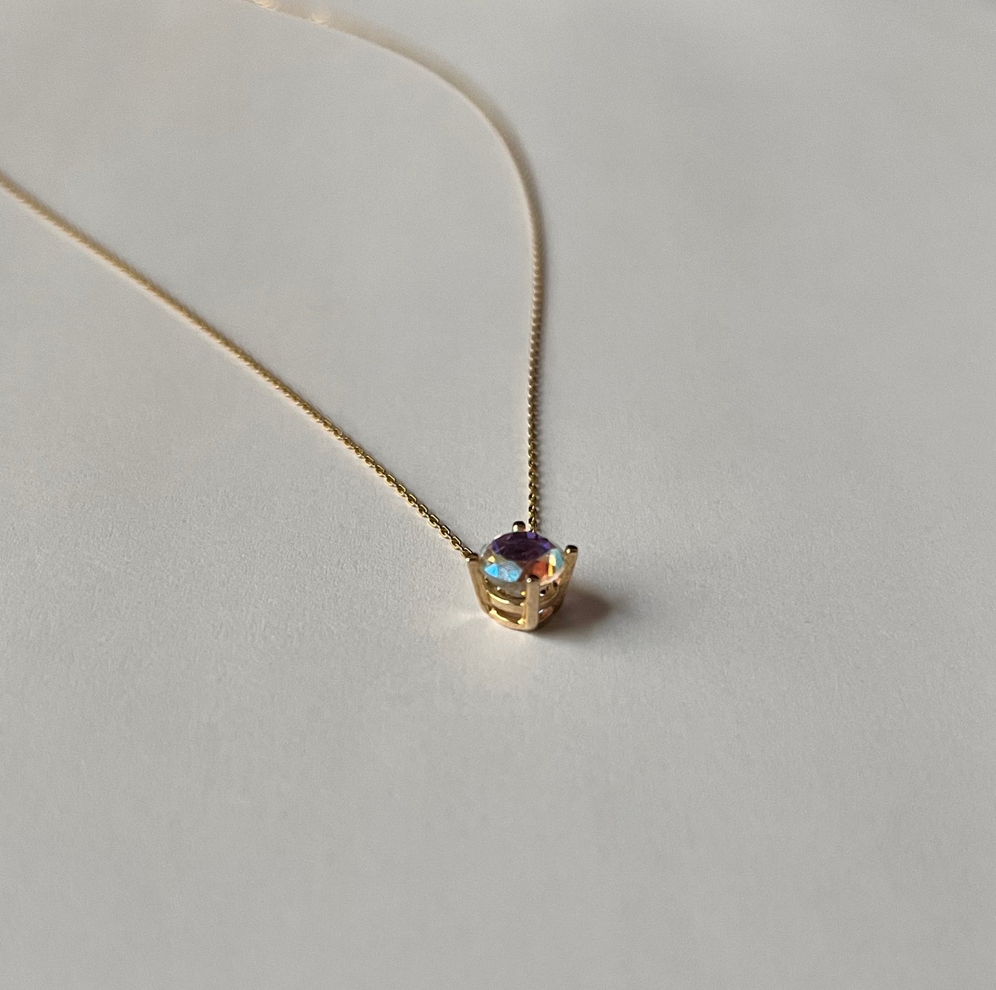 14k gold solitaire necklace with round mystic topaz pendant showing deep purple and blue rainbow flash, angled view on cream background