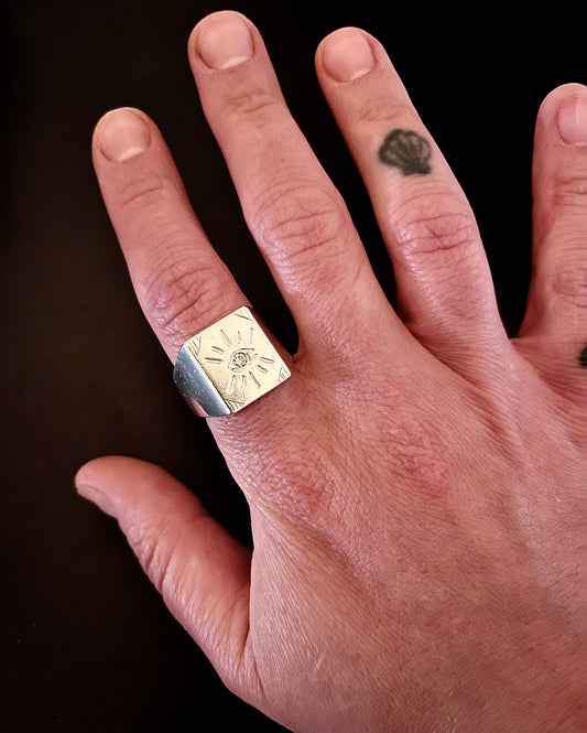 Sterling silver Eye of Protection signet ring worn on index finger, showing engraved evil eye motif, bold wide band, on tattooed hand against dark background