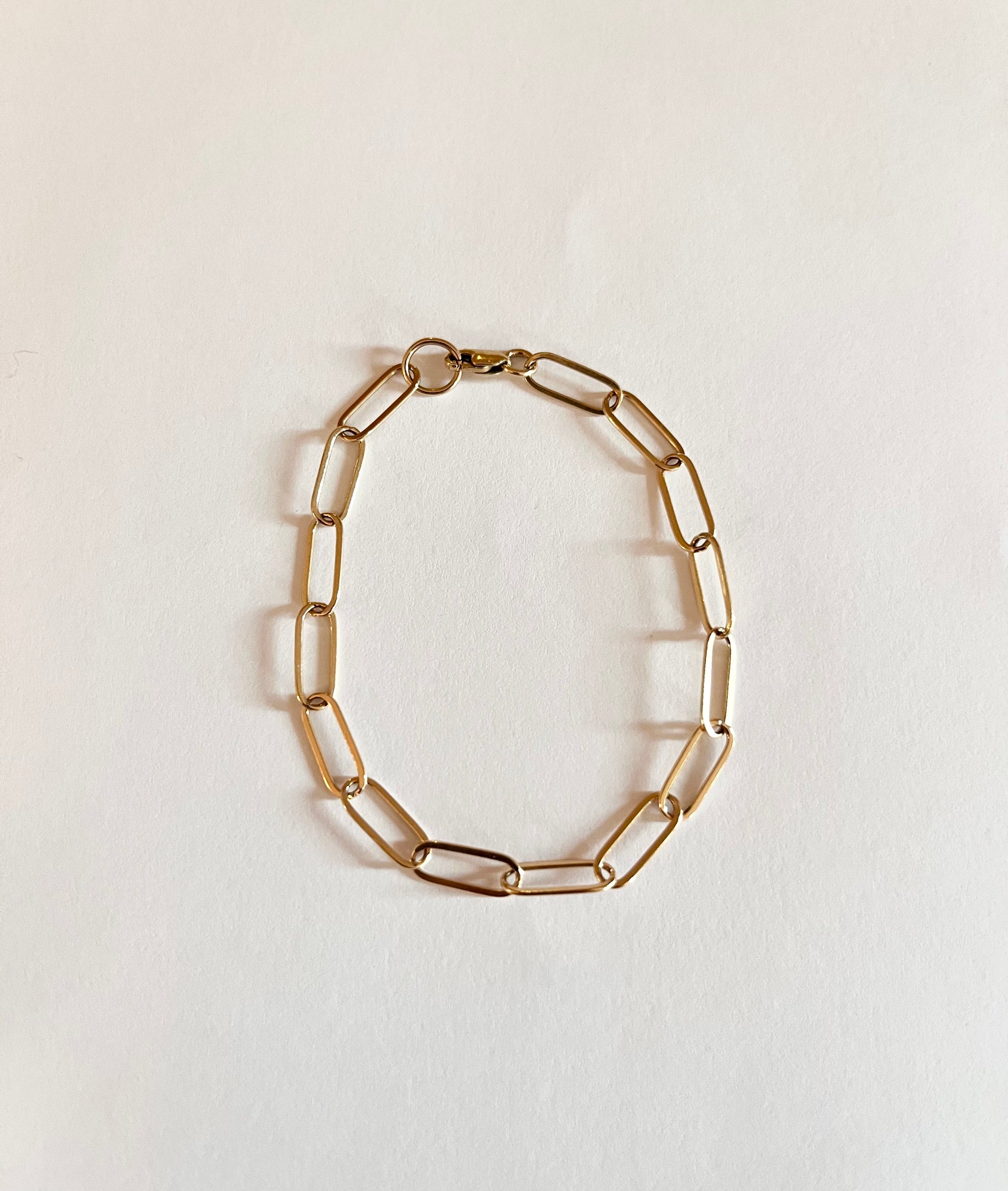 A gold paperclip chain bracelet laid out on a white surface.